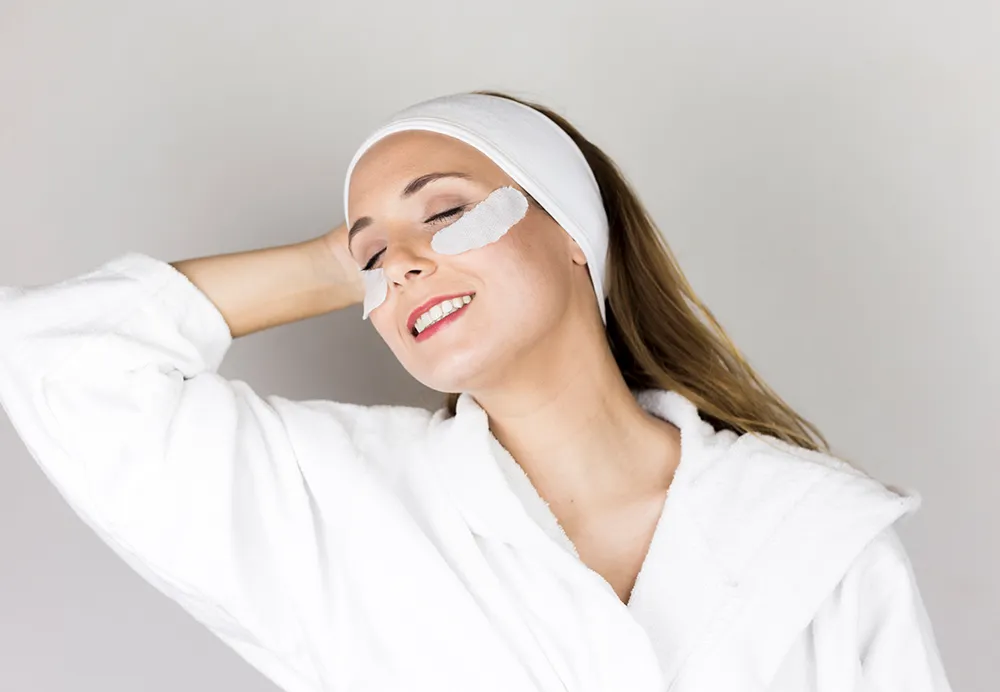 A smiling woman wearing a white headband and robe with treatment patches under her eyes Text What is EyeRevive Peel Treatment 1000x692